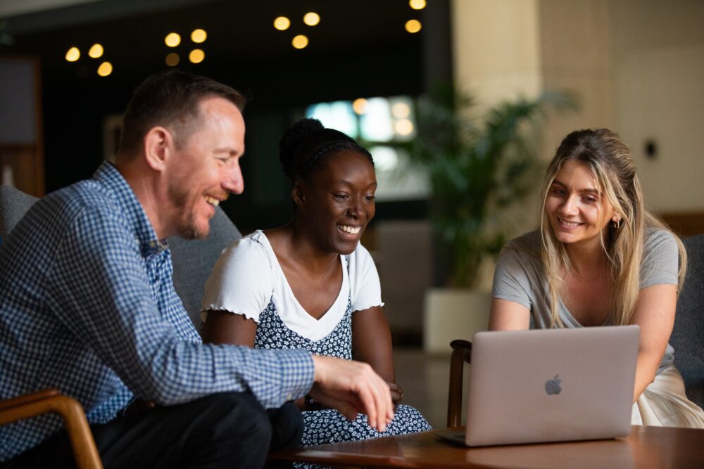 Three people sitting on chairs looking at a laptop placed on a table.